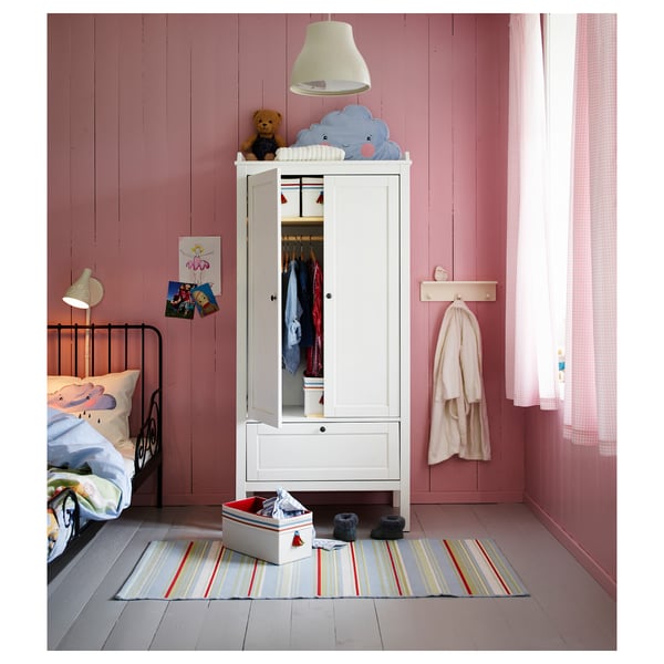 Kinderkamer met witte SUNDVIK kledingkast, beer, kleding, vloerkleed, roze muren, zwart bed, lamp, posters en gordijn.