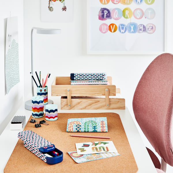 Een kleurrijke, kurken bureauonderlegger met verschillende kantoorartikelen zoals potloden, stickers en blocnotes, op een wit bureau met een rode stoel.