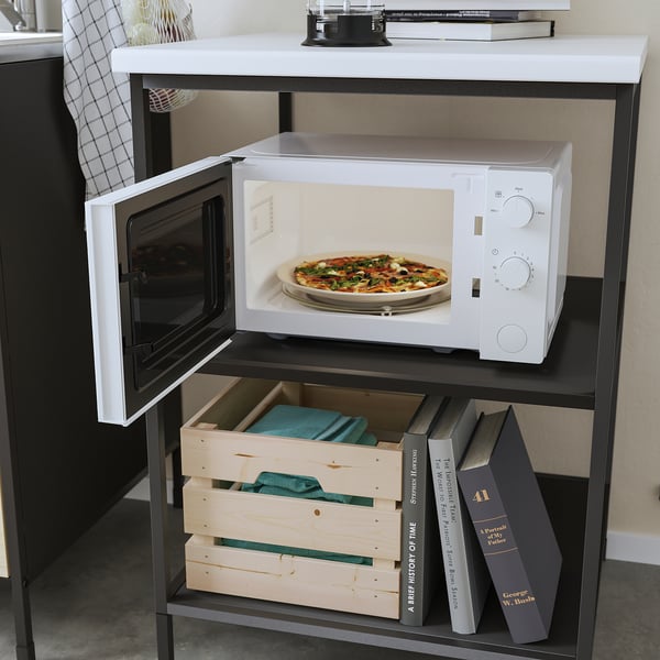 Een witte TILLREDA magnetron op een zwarte standaard, het opwarmen van een pizza. Beneden zijn er boeken en houten dienbladen.