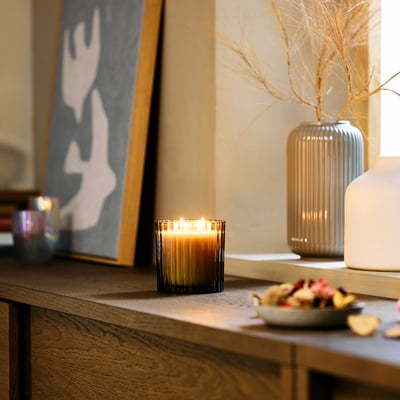 Een gezellige woonruimte met een TOFSTÄRNA geurkaars. Aangestoken kaars op een plank met decoratie, vazen en abstracte kunst.