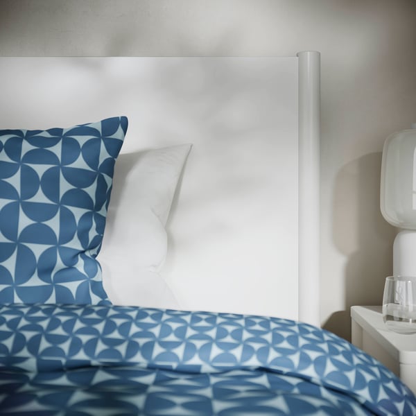 Bed met wit frame, blauw beddengoed met cirkelvormig patroon, witte kussens. Vacuüm toegankelijke ruimte onder het bed.