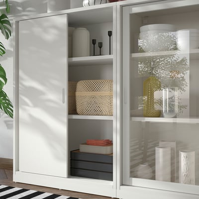 Witte TONSTAD opbergkast met schuifdeuren en glazen deuren, voorzien van gevlochten manden, vazen en dozen. Modern, gebroken wit ontwerp met strakke lijnen.