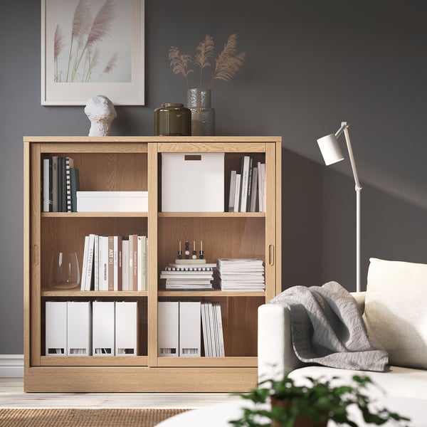 Gezellige woonkamer met TONSTAD eikenhouten boekenkast, gevuld met boeken, en een witte fauteuil, tegen donkere muren.