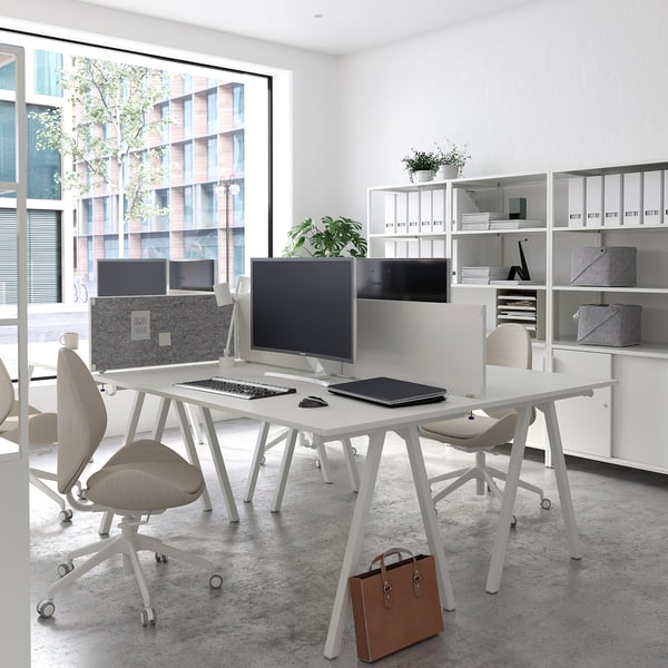 Modern kantoor met TROTTEN-bureaus, monitoren, beige stoelen, boekenplanken bij groot raam, uitzicht op de stad.