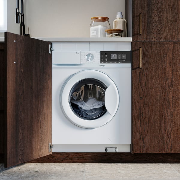 Een witte inbouwwasmachine genesteld tussen houten kasten met daarop een bedieningspaneel en wasmiddel.