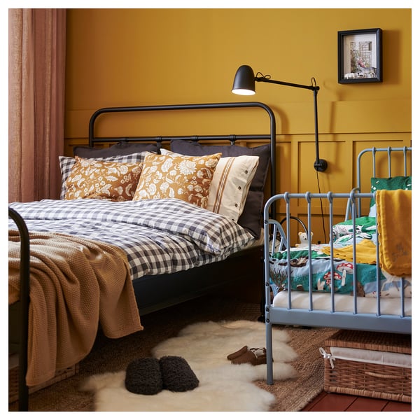 Gezellige slaapkamer met zwart bed, gele kussens, blauw kinderbed, beige vloerkleed, slippers, wandkandelaar, ingelijste foto op gele muren.
