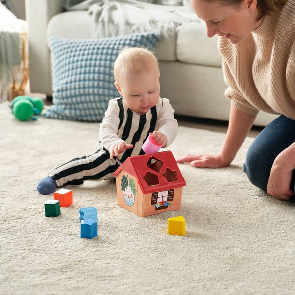 Baby spelen met vorm sorteerder speelgoed, geleid door volwassene. Het kind houdt roze blok dichtbij houten huisvormig stuk speelgoed met gekleurde blokken rond.