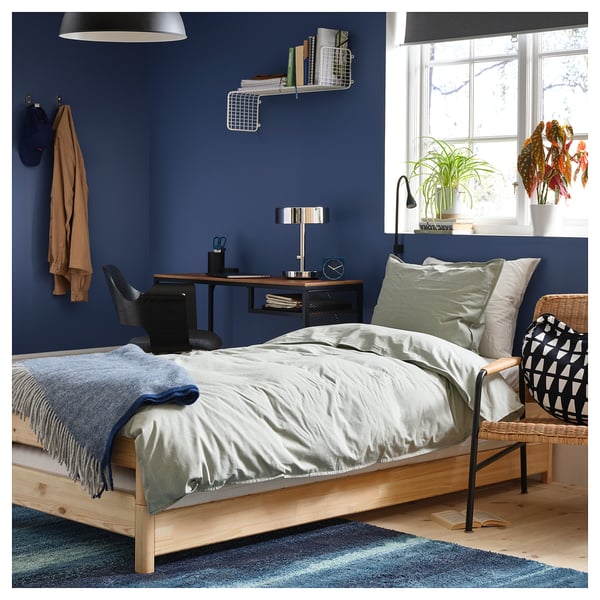 Gezellige blauwe slaapkamer met houten bed, wit beddengoed, groene kussens, zwarte stoel, bureau en raamplanten.