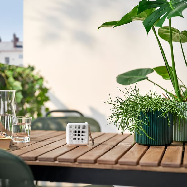 Kleine witte VAPPEBY-luidspreker zit op een houten tafel, omringd door planten en drinkglazen. Compact en draagbaar.