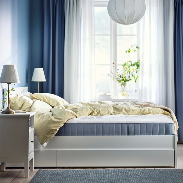 Slaapkamer met wit bed, lichtblauwe gordijnen en geel beddengoed. Twee witte lampen op nachtkastjes. Groot raam met hangplant.