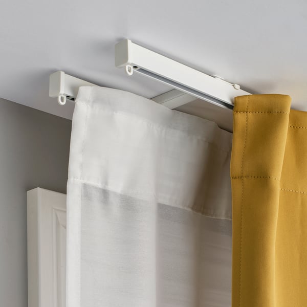 Witte plafondgemonteerde gordijnrails met twee lagen gordijnen. Doorschijnende witte binnen- en mosterdkleurige buitengordijnen hangen soepel aan zweefvliegtuigen.