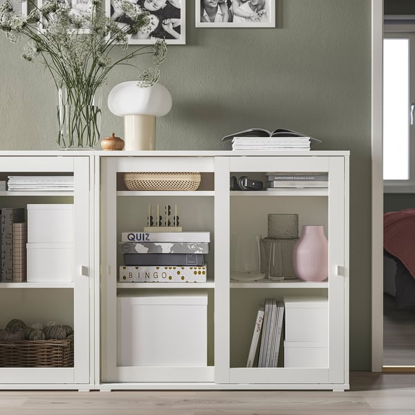 Moderne witte vitrinekast, kleiner dan omringende meubels, gestyled met vazen, boeken en decor.