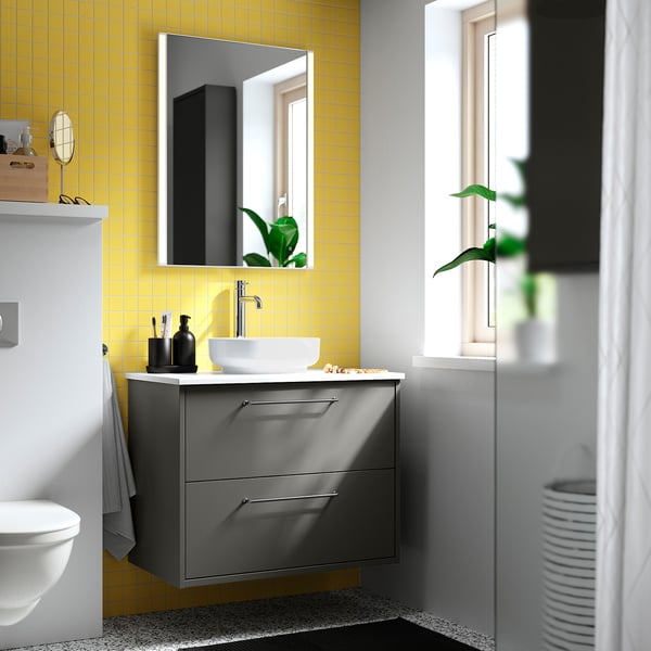 Licht badkamer met gele tegels, witte wastafel op een grijs kastje, grote spiegel, kleine plant en toilet. Grijze vloer op de vloer.