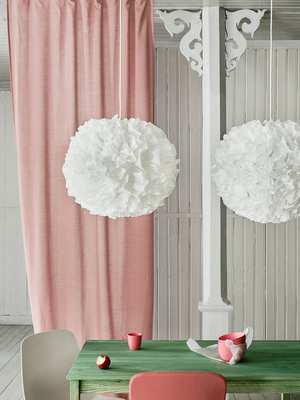 Boven een groene tafel met een rode mok en appel hangen twee witte bolvormige hanglampen. Roze gordijnen aan de linkerkant.