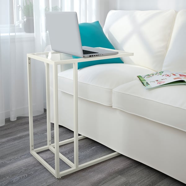Een moderne woonkamer, een witte VITTSJÖ laptopstandaard met een glazen blad, een opengeslagen laptop naast een witte bank met blauwe kussens.