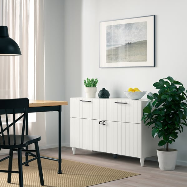 Scandinavian-style dining area, featuring BESTÅ white cabinet with black handles, positioned between dark wood chairs and a large potted plant.