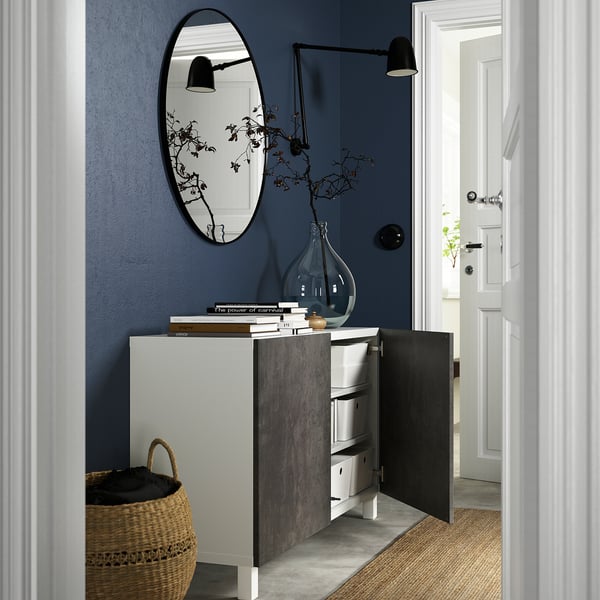 Small entryway with BESTÅ sideboard, open door revealing storage. Dark blue wall, mirror, black switch.