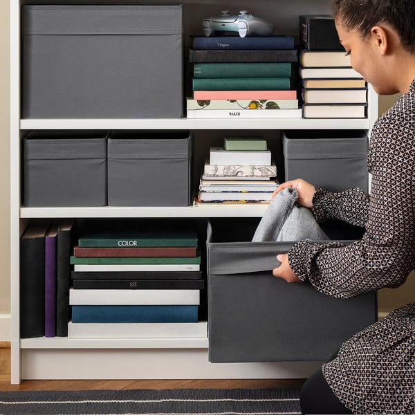 Person organising grey fabric storage boxes on white shelves, holding one box in hand, surrounded by books and gaming controllers.