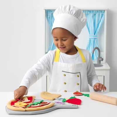 Child wearing chef hat and apron playing with felt pizza toys on a light-colored countertop.