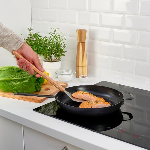 Person grilling fish in a pan on a modern stove. Kitchen tools and greens visible.