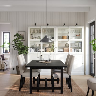 Modern dining room featuring HAVSTA white cabinets with glass doors, black metal table, grey upholstered chairs, and a large potted plant.