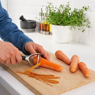 Person peeling carrots on a wooden cutting board with a metal peeler. Carrot shavings are visible on the board.