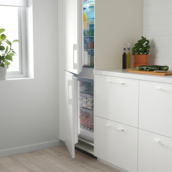 Open white fridge freezer reveals shelves with food. Next to it, a countertop with bottles and a potted plant.