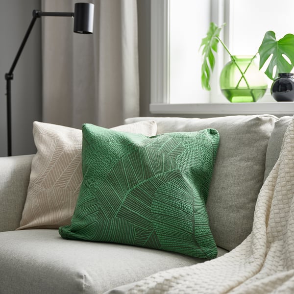 Green leaf-patterned cushion on couch with other pillows, a blanket, lamp, and potted plants.