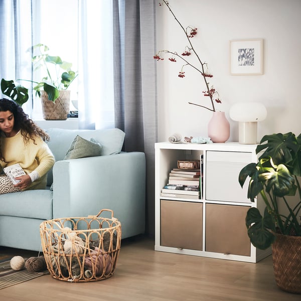 Cosy living room: person reads on blue KALLAX sofa next to shelves with books and vases, yarn basket on floor.