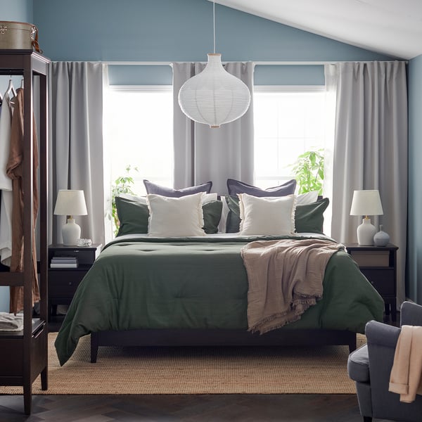 Green comforter bed, lantern, grey curtains, nightstands, lamps, rack, rug.