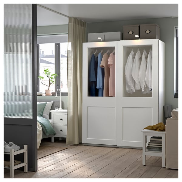 Modern bedroom with white PAX wardrobe showing clothes, a bed, bedside table, and window with curtains.