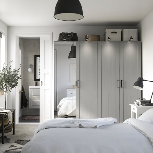 Modern bedroom with grey PAX wardrobe, mirrored door, and glass panels. Decor includes plant, lamp, and bed with white linens.