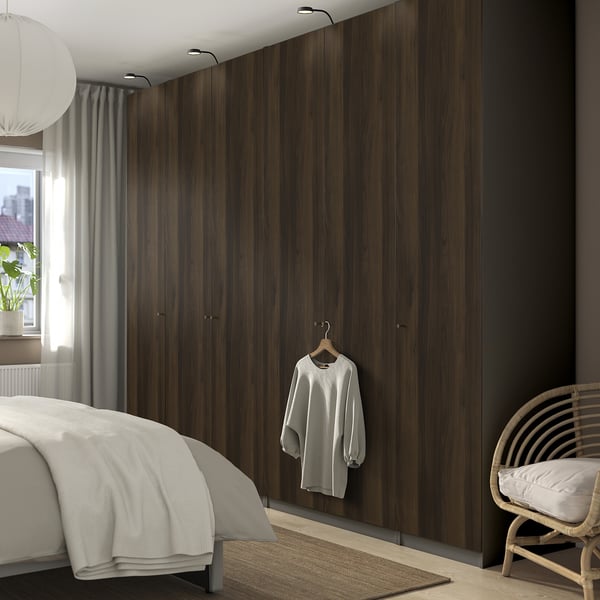 Modern bedroom with tall dark wood PAX wardrobe, white bed, wicker chair, and large window.