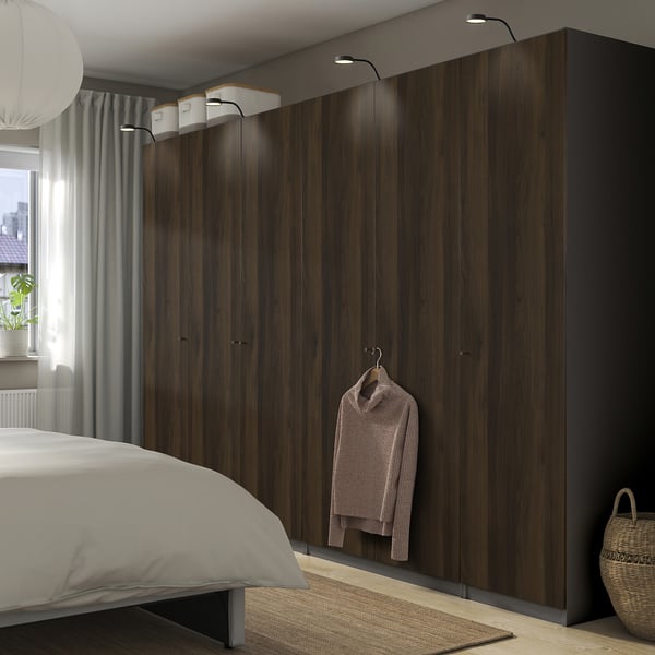 Modern bedroom with large PAX wardrobe featuring dark wood grain, integrated lights, hanging garment, white bed, grey rug, and wicker basket corner.