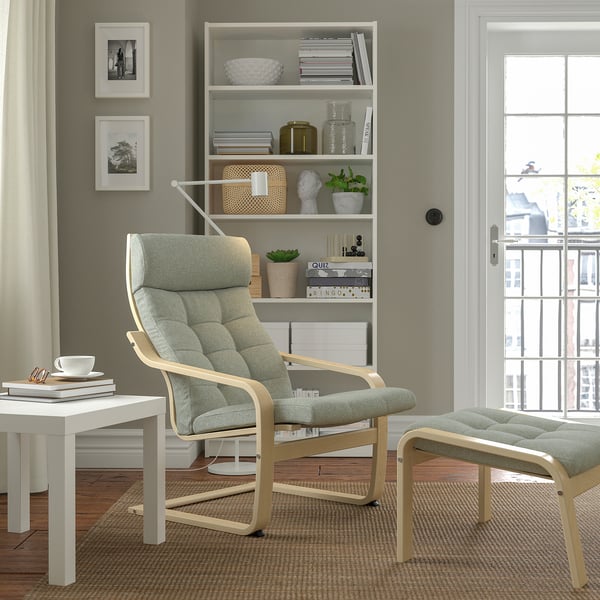 Modern living room with light grey POÄNG armchair, ottoman, and shelves. Neutral decor, blackout curtains, wooden floor.