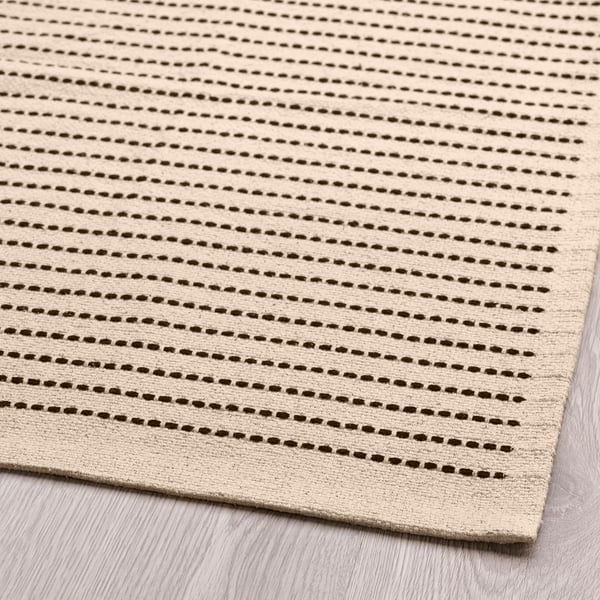 Beige rug with brown stripes, neatly laid on wooden floor, showcasing high-quality cotton texture and even stitching.