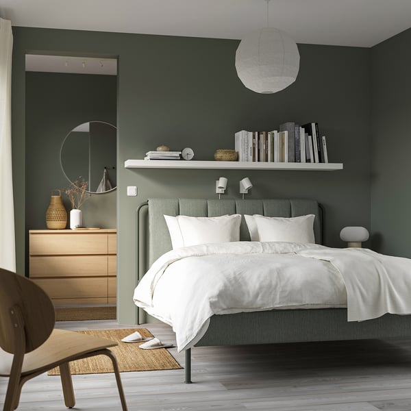 Modern minimalist bedroom with TÄLLÅSEN bed in gray-green. White linens, wooden dresser, and round mirror. Bookshelf and woven chair.