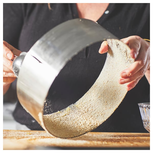 A person uses a stainless steel, round, adjustable baking frame on a wooden surface to bake or cut a cake.