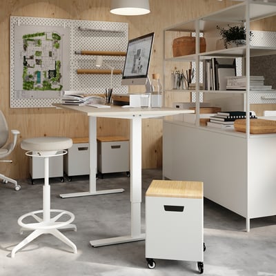 A modern office setup with a standing desk, white TROTTEN with a beige top, and an adjustable stool. It includes shelves with books and decor.