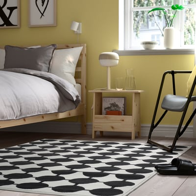 A modern bedroom with a black & white VÄNDPLATS rug, wooden bed, grey/white bedding, yellow wall, and minimalist decor.
