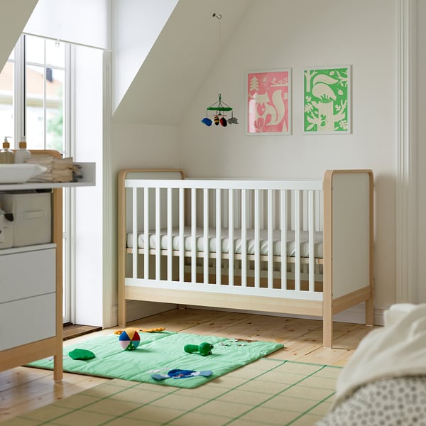 Minimalist nursery with ÄLSKVÄRD cot, light wood, colourful art, changing table, and green mat.