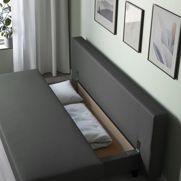 Modern sofa with hidden bed; white pillows, grey upholstery; pictures on wall; storage under seat.
