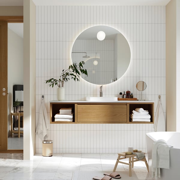 Minimalist bathroom with round mirror, light wood drawers, greenery, open design.