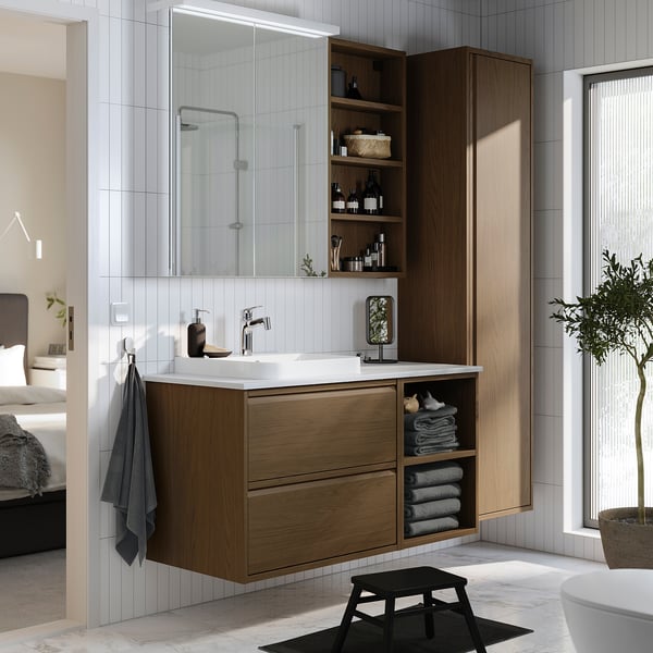 Modern bathroom with ÄNGSJÖN oak vanity, white sink, large mirror, and open shelves. Towels on the side, black stool, and houseplant.