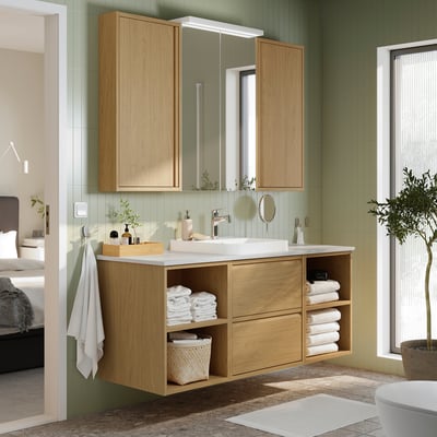 Modern bathroom vanity with marble top, green tiles, and mirror lights, plant nearby, white bedding in adjacent bedroom.