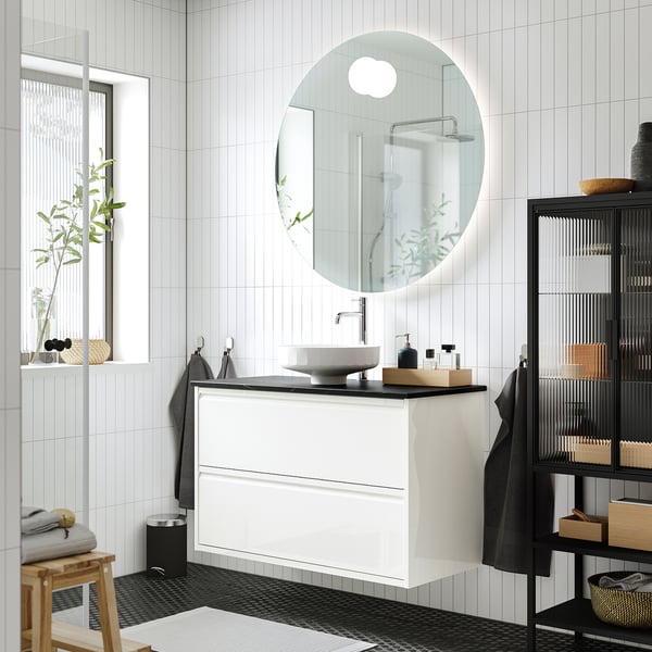 Modern bathroom with ÄNGSJÖN washstand, black vanity, and storage. Circular mirror reflects minimalist decor, wooden stool with plant.