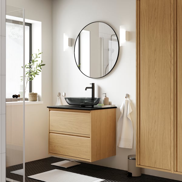 Modern bathroom with ÄNGSJÖN light oak vanity, dark glass sink, round mirror, and towel hooks. Compact yet spacious design with minimalist style.