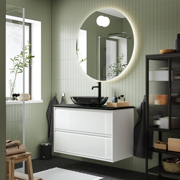 Modern bathroom with white ÄNGSJÖN vanity, black sink, and round mirror. Green tiled walls, wooden accents, and black fixtures.