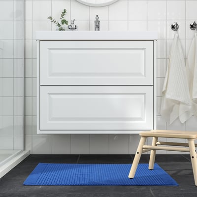 Modern bathroom with white cabinet, glass shower, blue ALSTERN mat, wooden bench, and white towels.