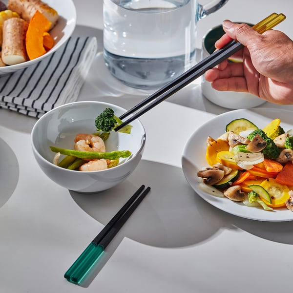 Hand holds coloured chopsticks near bowls of veggies and shrimp. Square tips prevent rolling.